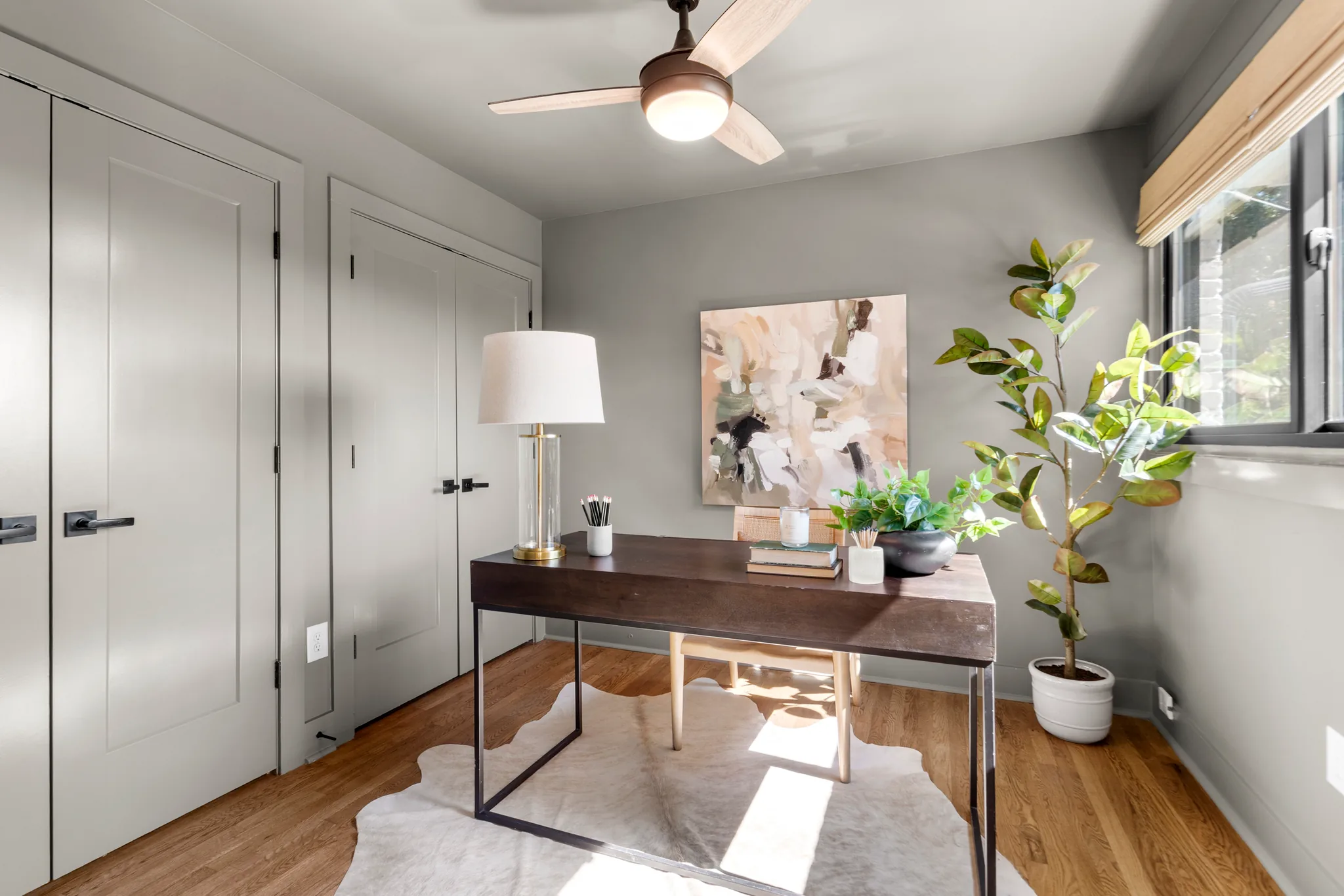 Small home study with gray walls, white closet doors with black hardware, dark wood desk on a hide rug and oak floors