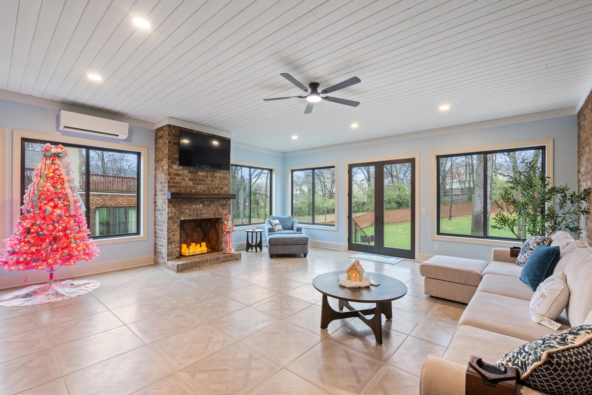 Enclosed sunroom with brick fireplace and wall-mounted TV, white shiplap ceiling, ceiling fan, tile floor and windows overlooking a fenced yard