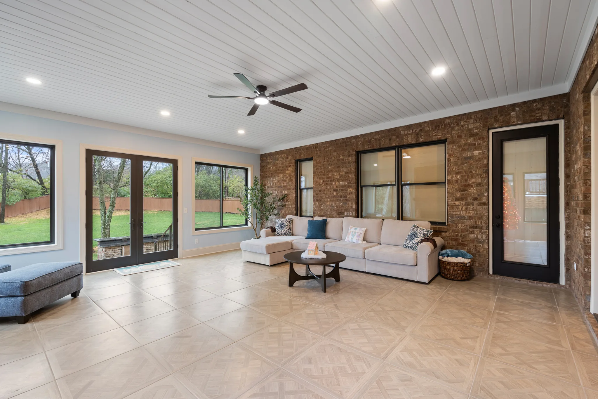 Enclosed sunroom with exposed brick wall, white shiplap ceiling, large tile floor, sectional sofa and French doors to a fenced backyard