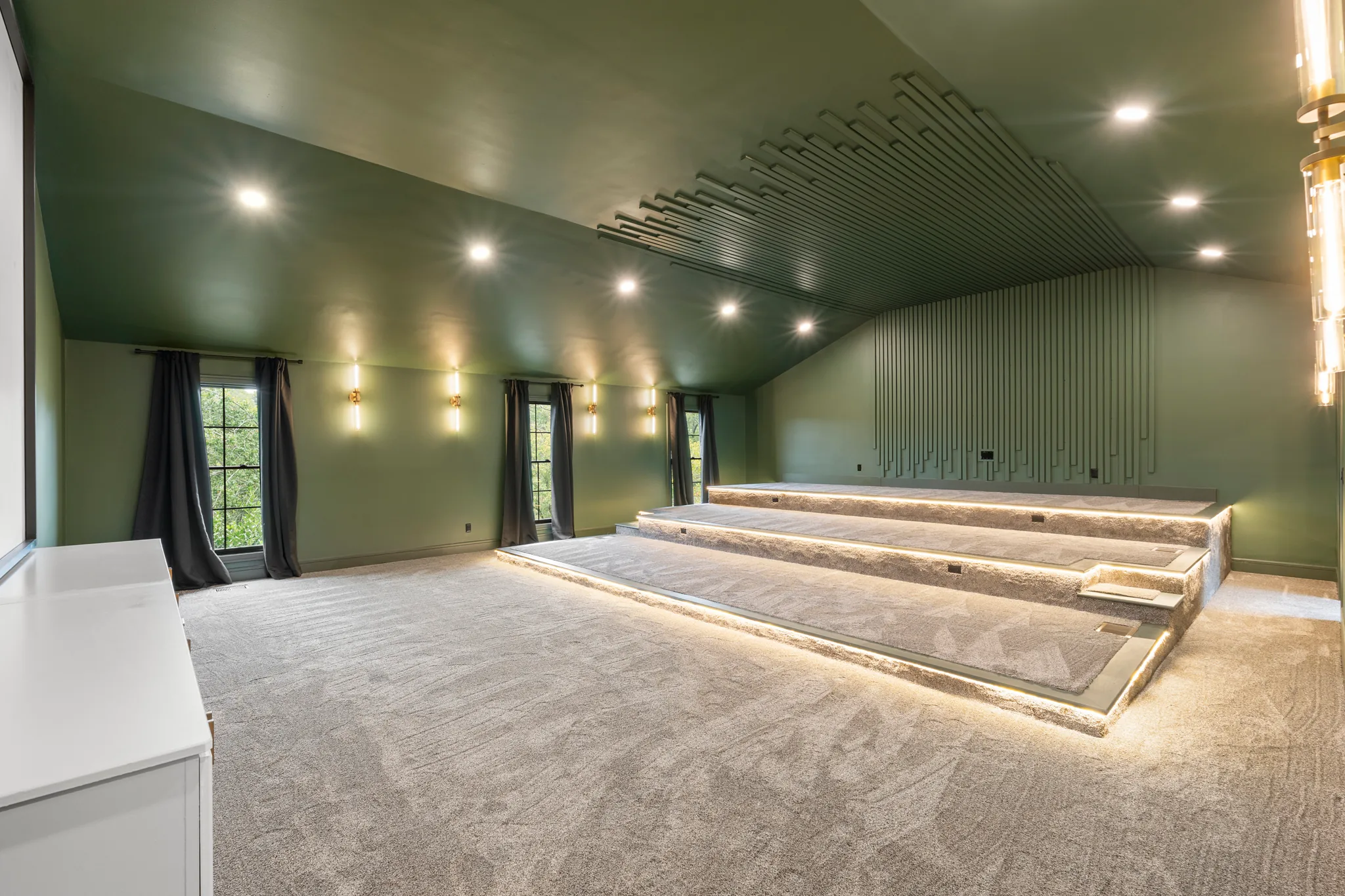 Home theater room with sage green walls and vaulted ceiling, carpeted tiered seating platforms lit by LED strips, and slatted wood wall treatment