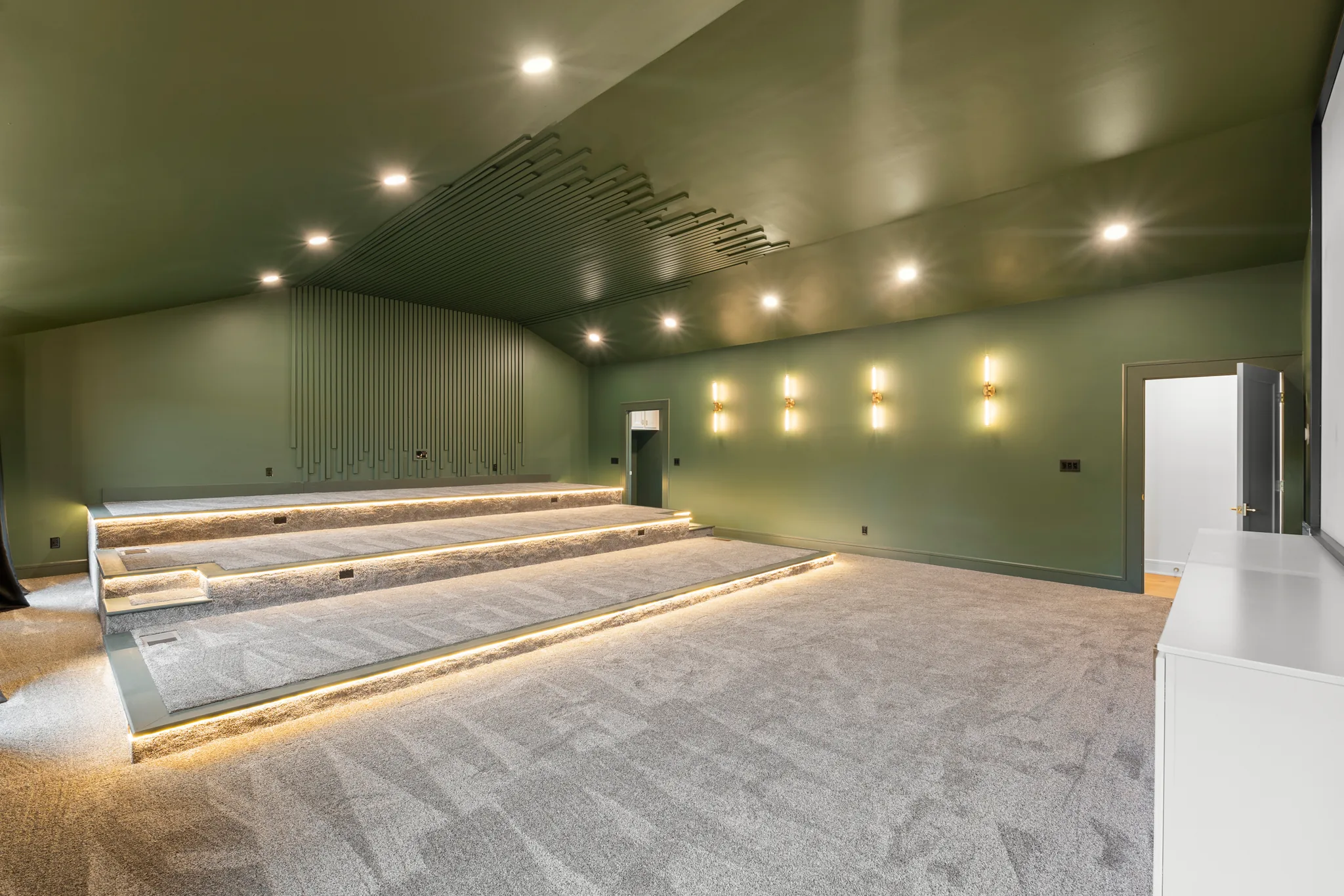 Home theater room with three carpeted seating tiers lit by LED strips, sage green walls, slatted wood accent panel and wall sconces