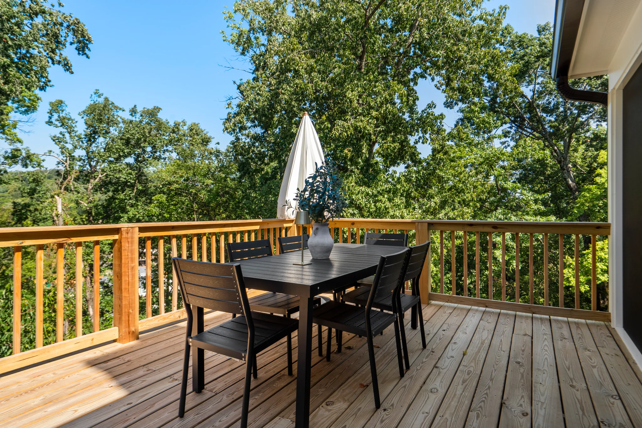 Wood deck with light natural pine decking and matching picket railing, holding a black dining set and closed white umbrella above green treetops