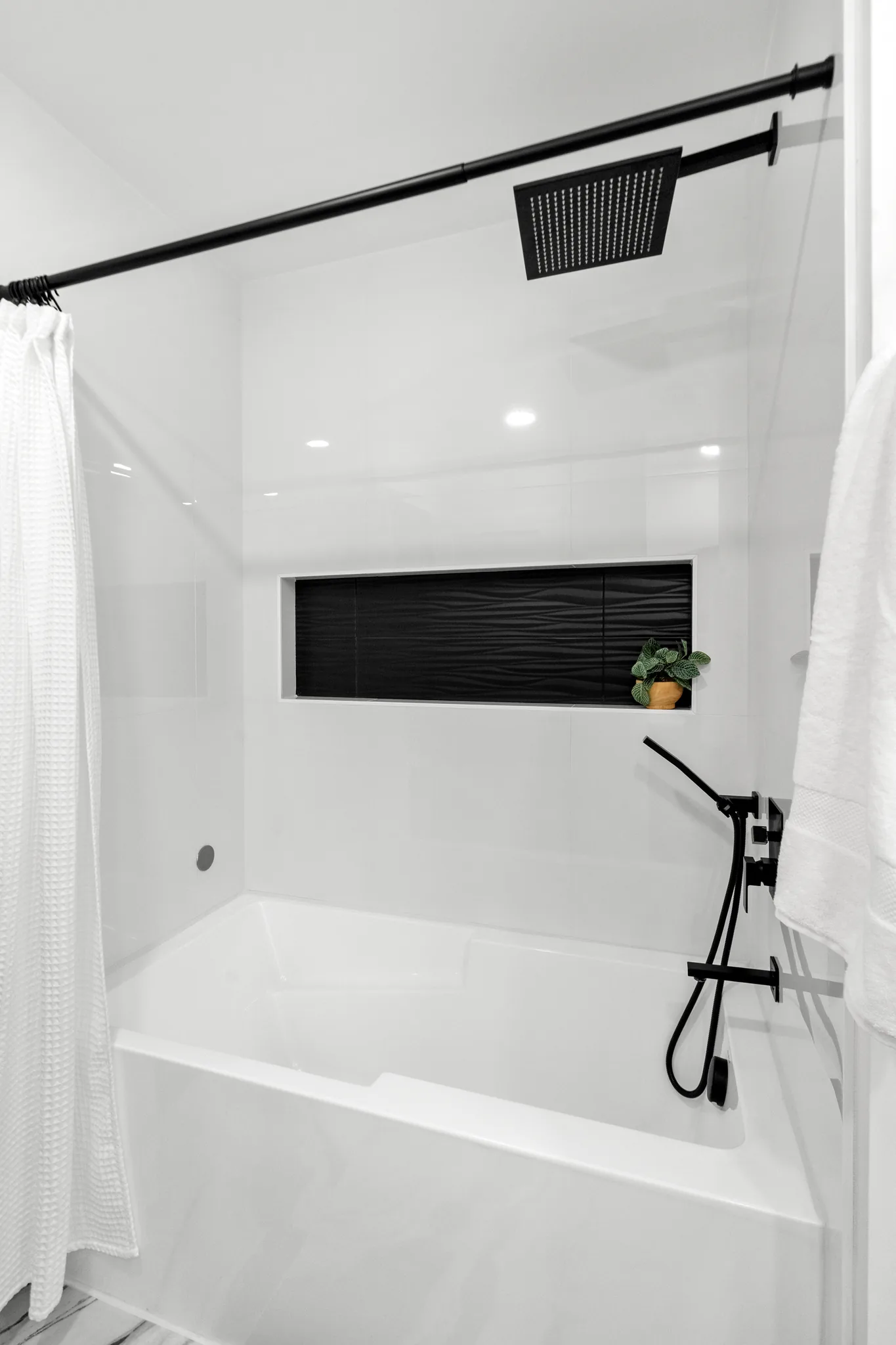 White tile tub and shower surround with a square black rainfall showerhead, black fixtures, recessed niche in black tile and a white waffle curtain