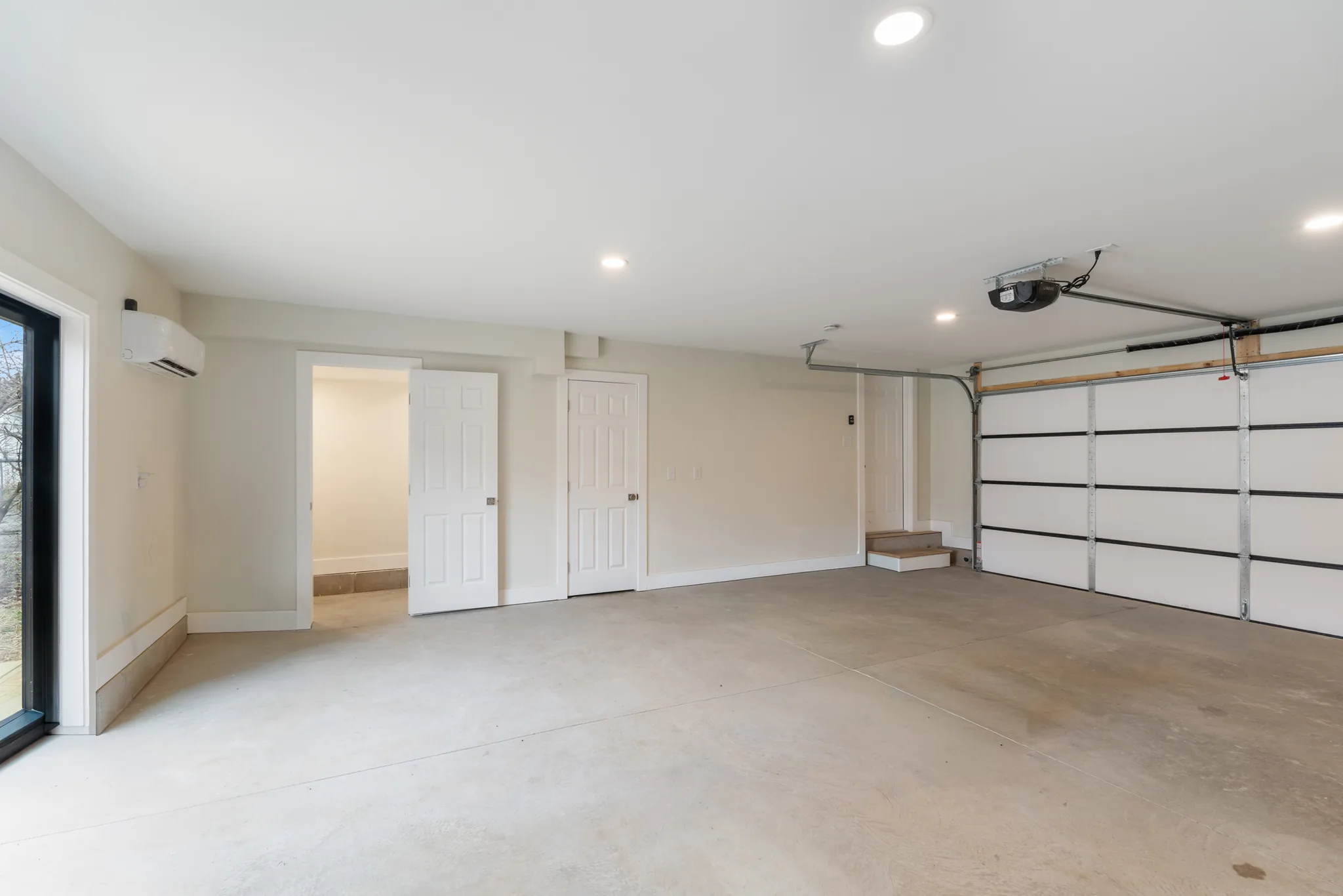 Empty finished two-car garage with smooth concrete floor, white sectional door, wall-mounted mini-split unit, recessed lights and two interior doors