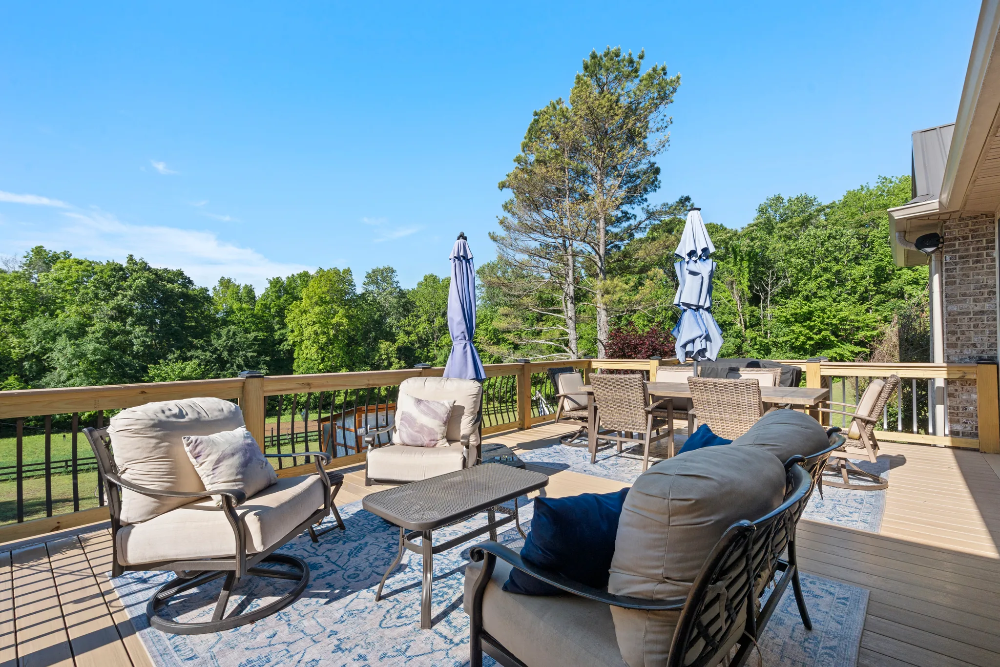 Two-level wood and composite deck with cushioned metal lounge chairs on a patterned outdoor rug, black balusters, closed umbrellas, and woods beyond