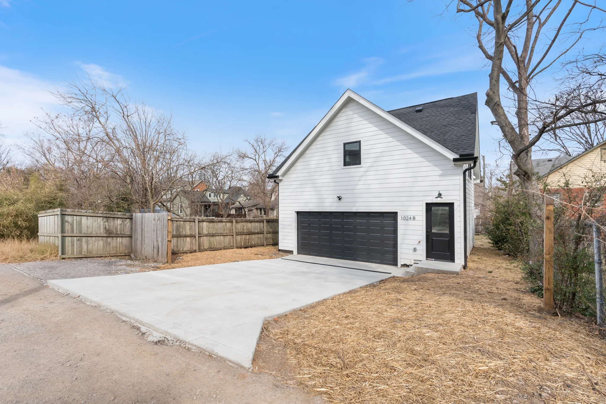 Two-story detached building with white lap siding, black sectional garage door, black shingled roof and a new concrete driveway beside a wood fence