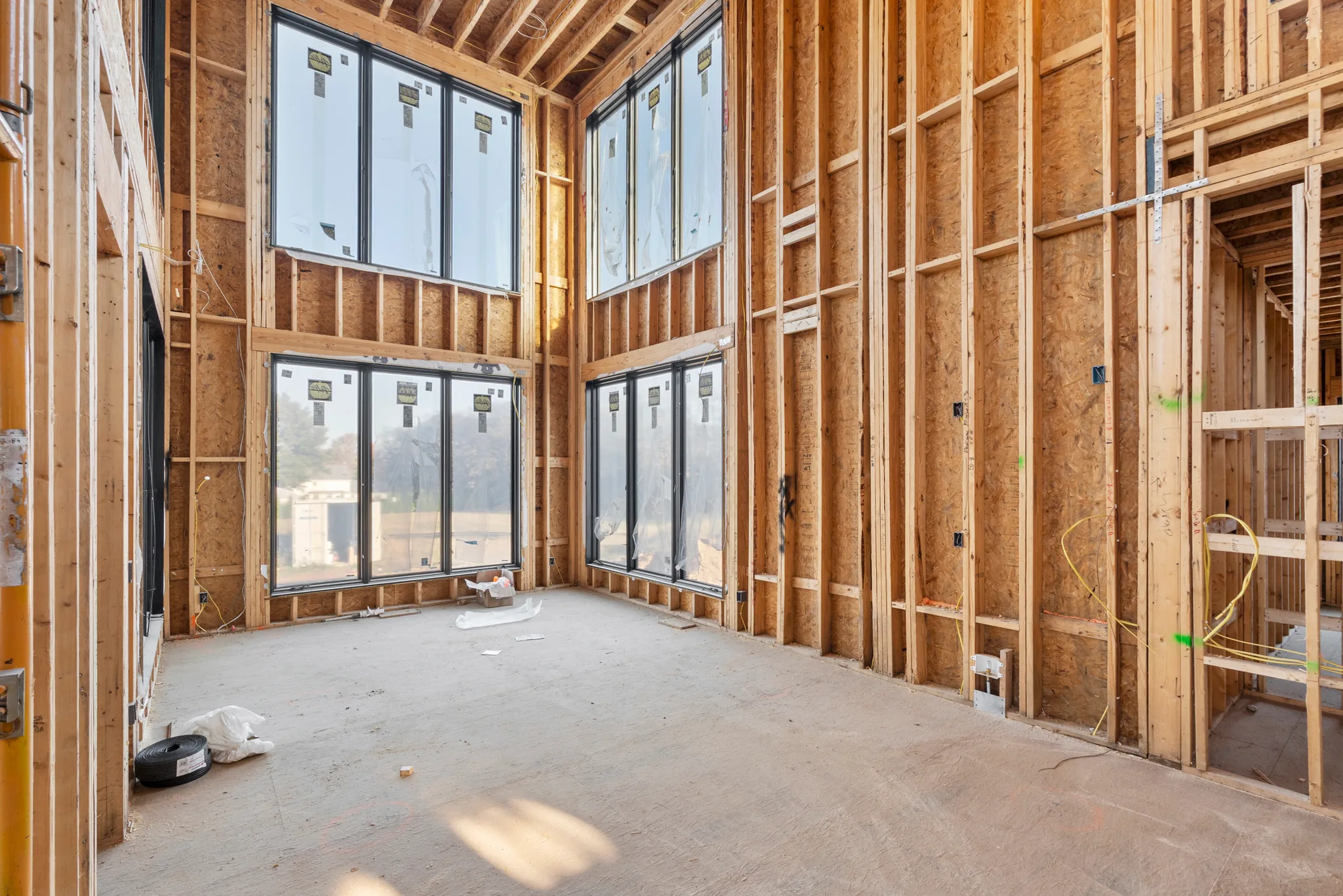 Interior framing under construction with stacked black window units, exposed studs, plywood sheathing and a plywood subfloor in a two-story room
