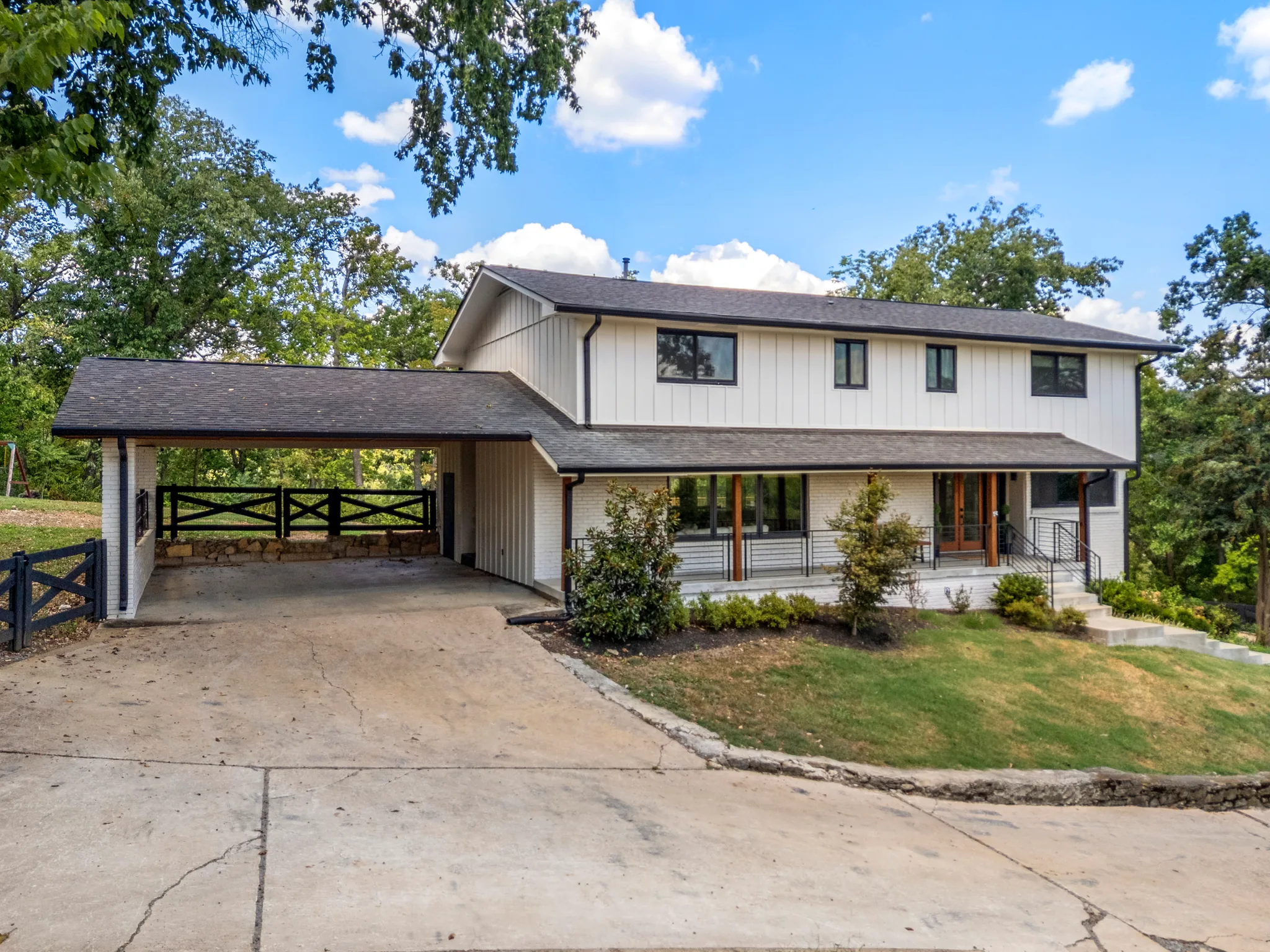 Two-story home with white board and batten upper level, painted brick lower level, black windows, covered front porch and an open carport reached by a wide concrete drive