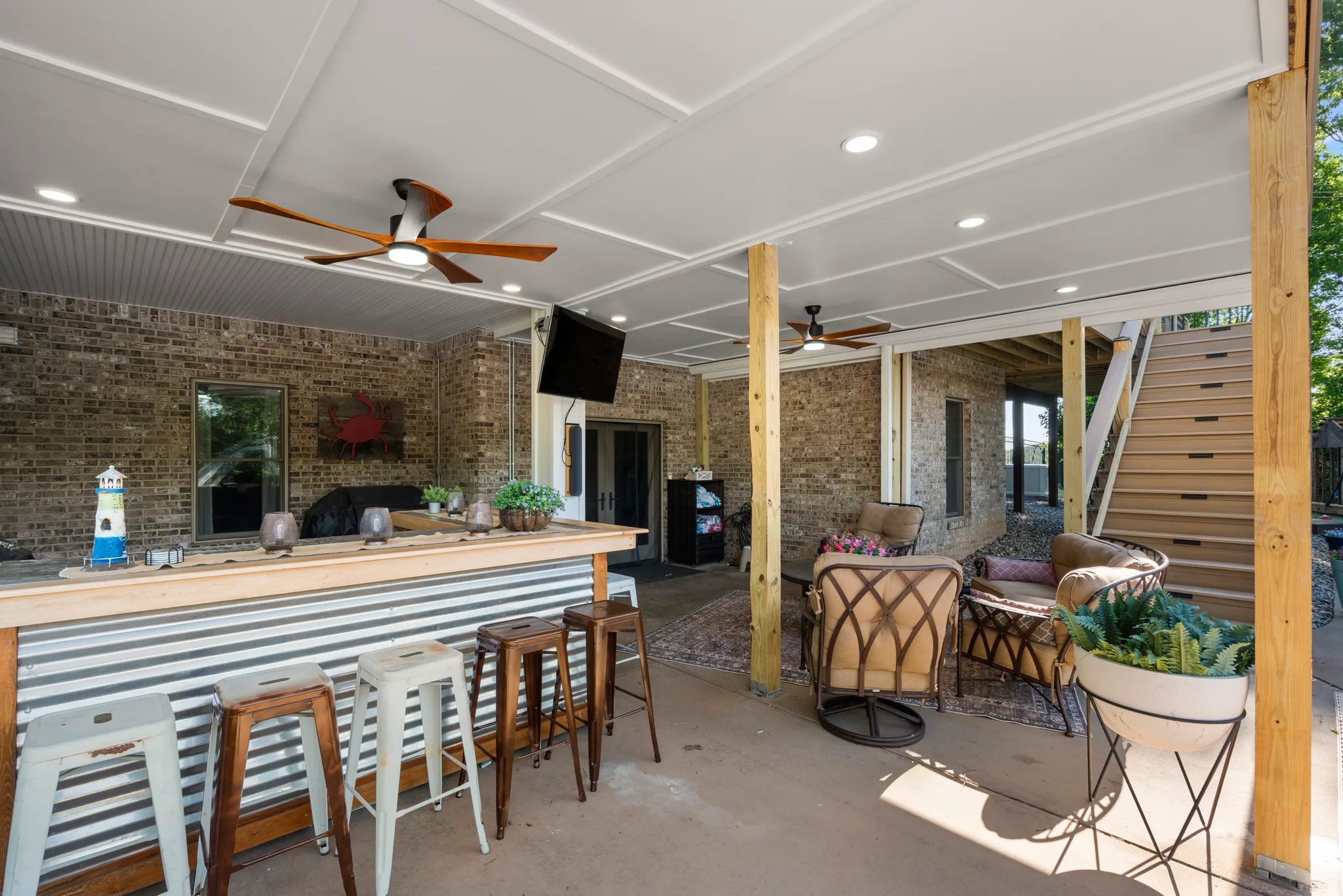 Covered outdoor bar with corrugated metal base, wood counter and mixed stools beneath a white paneled ceiling with fans, beside brick walls