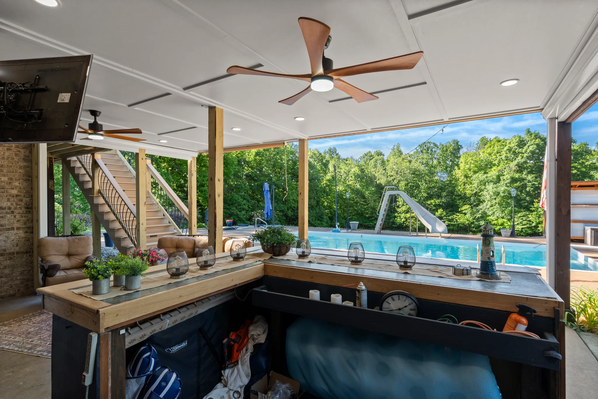 Covered under-deck bar with an L-shaped wood counter, ceiling fan and recessed lights, looking out to a pool with a slide and woods