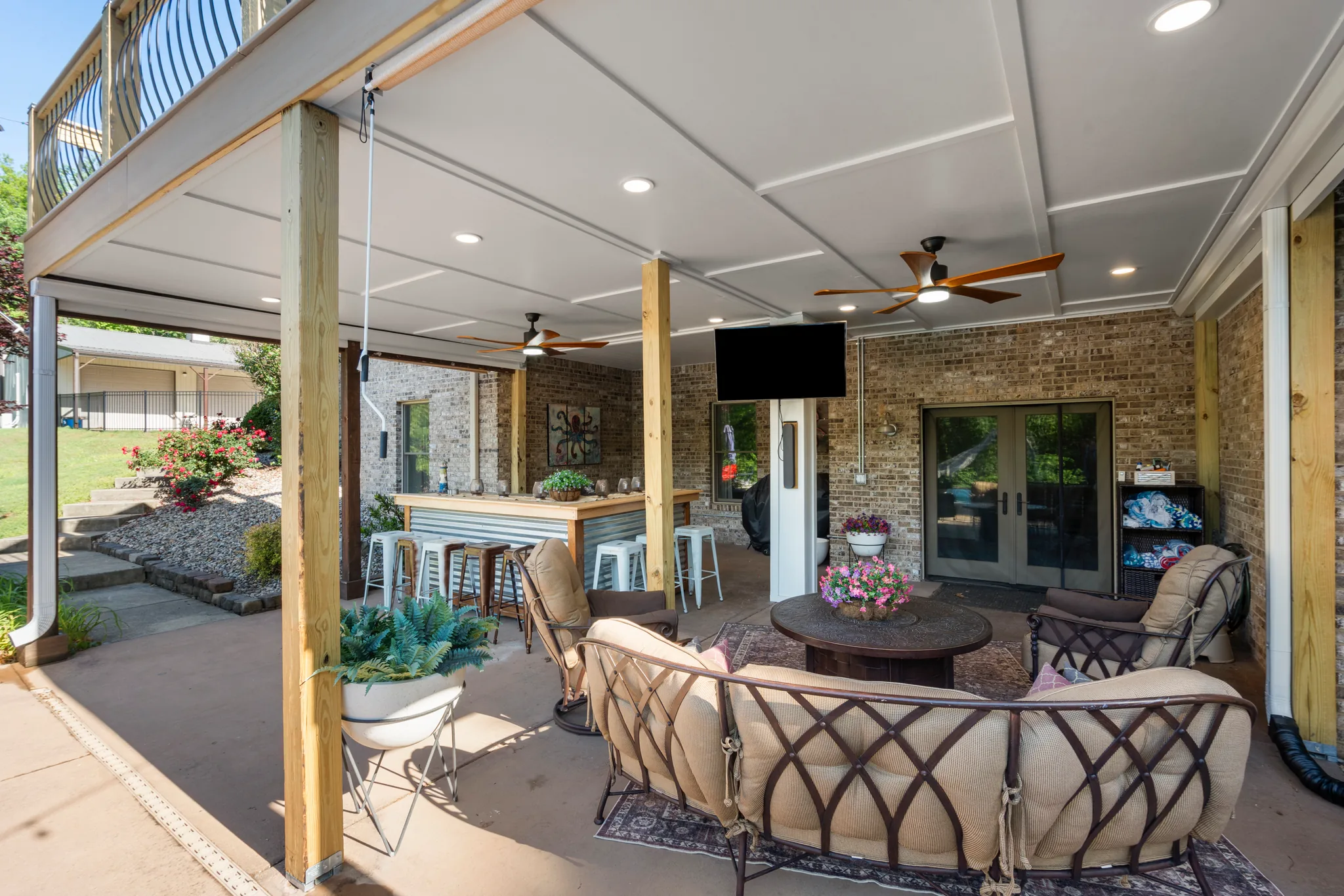 Covered patio beneath an elevated deck with white paneled ceiling, recessed lights, two fans, wall-mounted TV, wood bar with stools and cushioned seating
