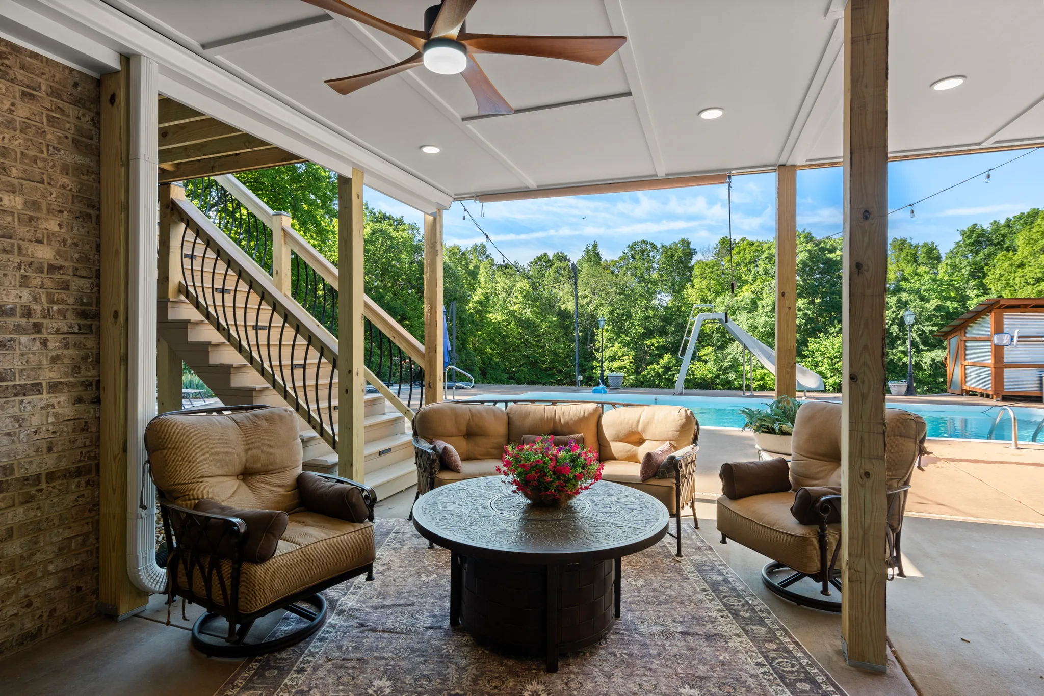 Covered patio under a deck with cushioned swivel chairs, round fire table, area rug and stairs leading up beside a brick wall overlooking a pool