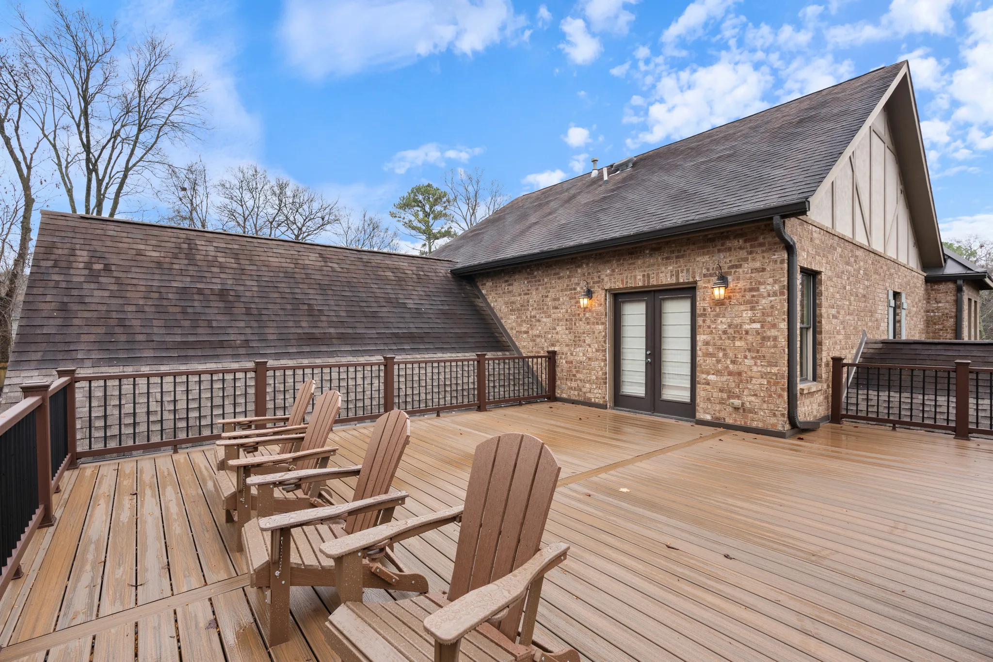 Wide wood deck with Adirondack chairs beside a brick house wall with black French doors, dark railing and shingled roof slope