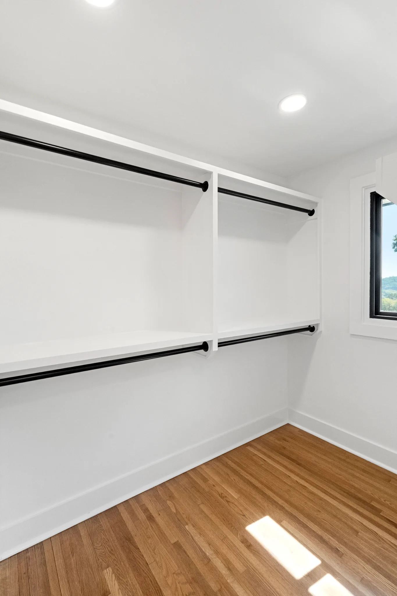 Empty walk-in closet with white shelves, black hanging rods, white walls, oak floors and a black-framed window in the corner