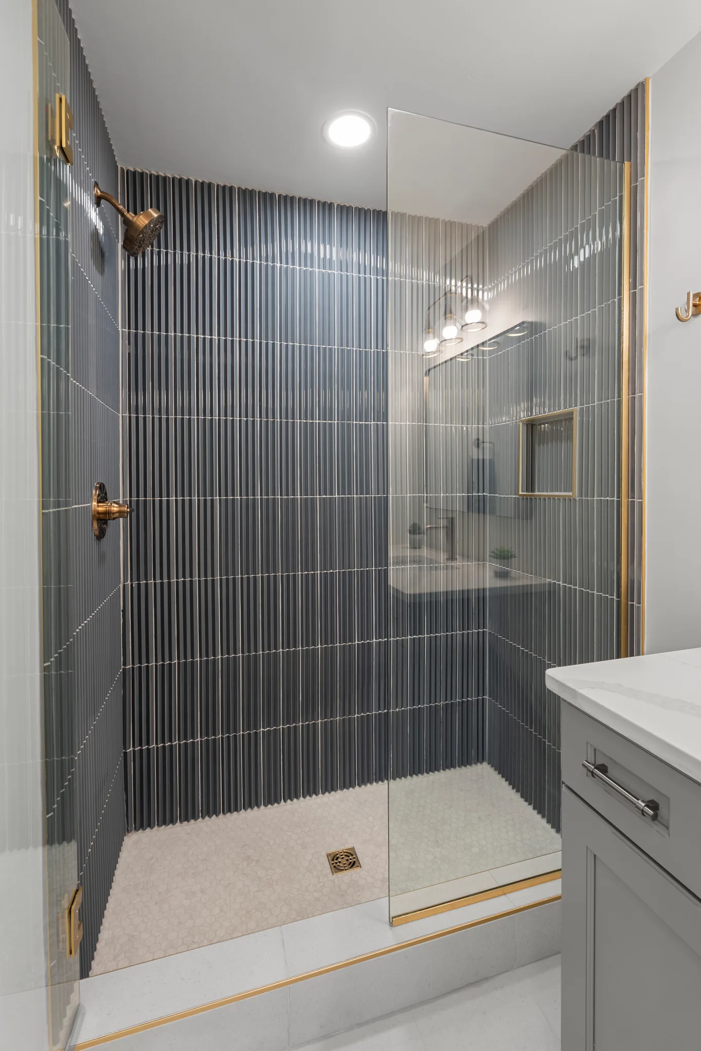 Walk-in shower lined with vertical fluted gray tile, brass shower head and valve, glass panel, pebble-look floor and gold trim edges