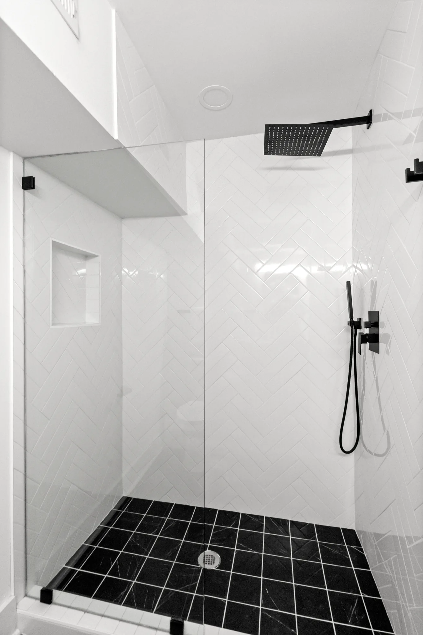 Walk-in shower with white herringbone wall tile, black marble floor tile, glass panel, matte black rain head and handheld sprayer