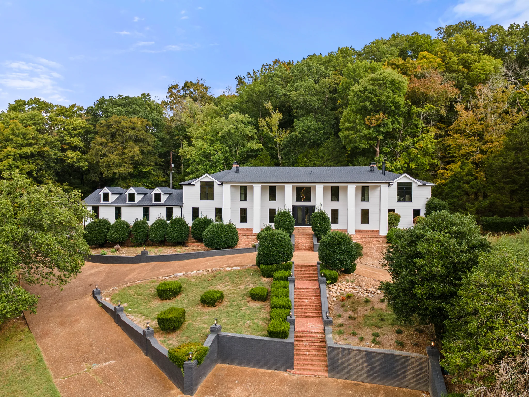 Aerial view of a long white two-story home with square porch columns, dark shingle roof, dormers and a brick stair climbing between painted retaining walls