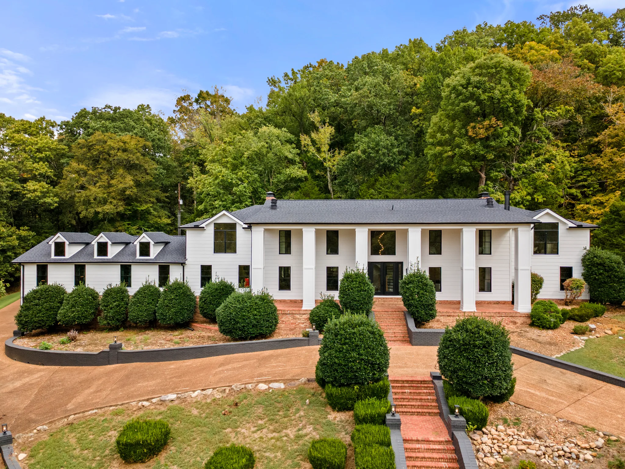 Elevated view of a long white two-story home with square columns, dark shingle roof, dormered side wing and brick front steps framed by clipped shrubs