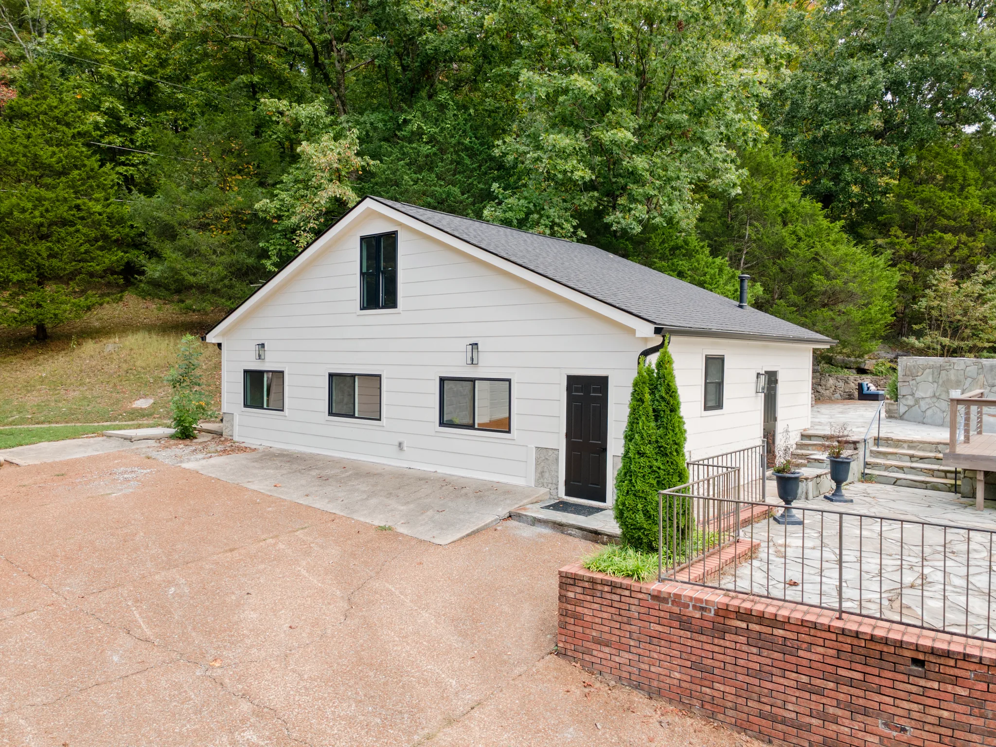 Small white lap-sided cottage with dark shingle roof, black door and black-framed windows beside a flagstone patio bordered by a brick retaining wall