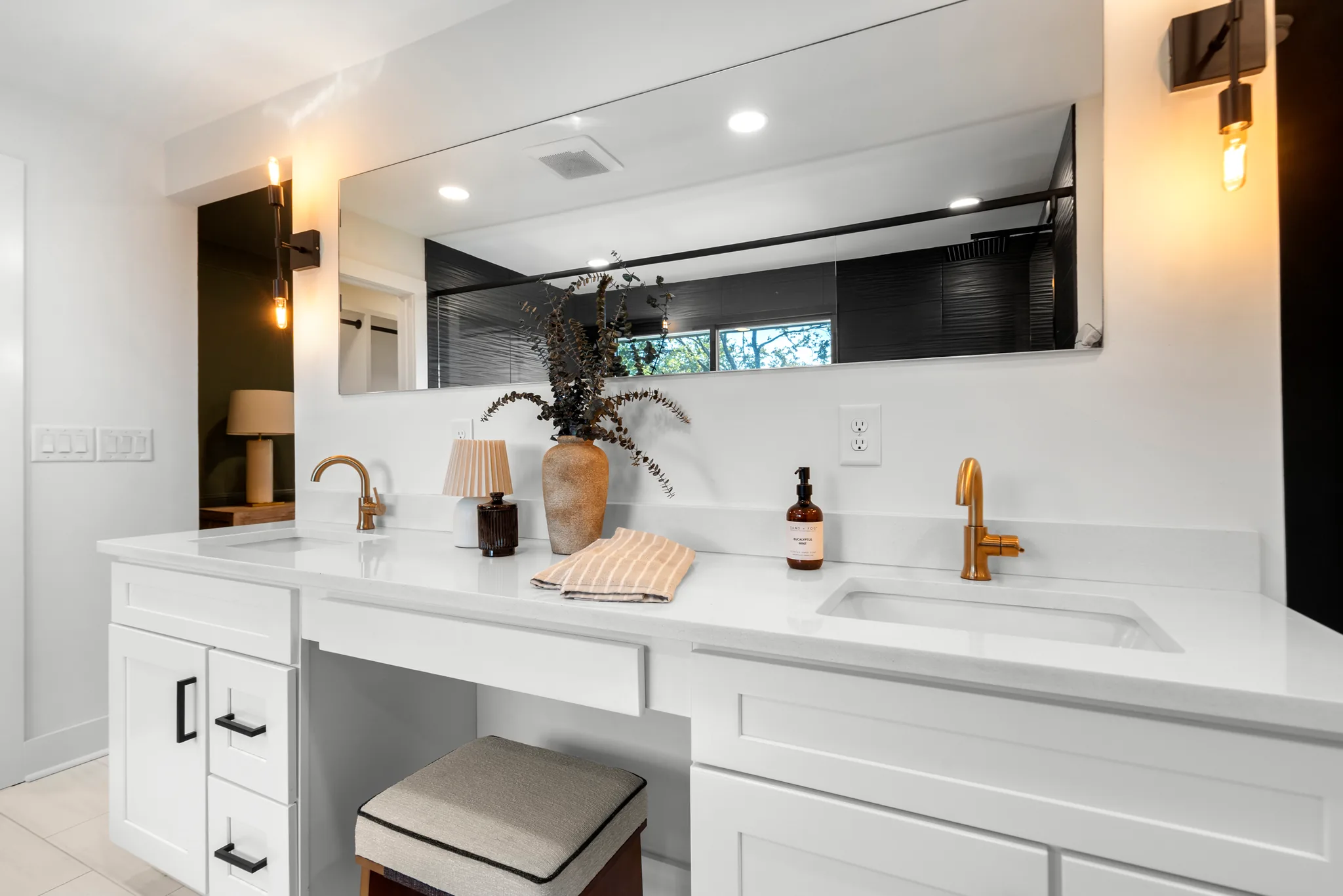 White shaker double vanity with quartz counter, brass faucets, center makeup station with upholstered stool, and a long frameless mirror reflecting a black tile shower