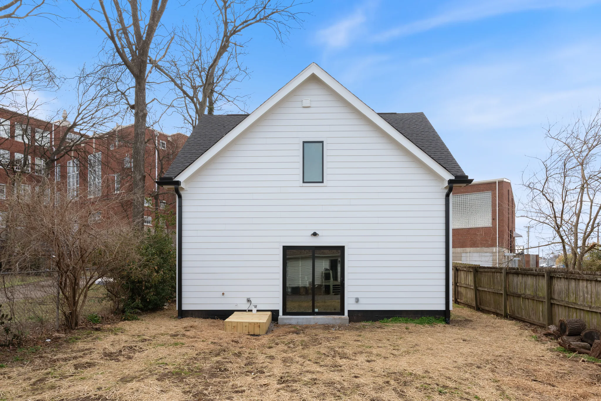 Rear elevation of a white lap-sided home with a steep gable, black-trimmed sliding glass door, tall narrow upper window and dirt yard