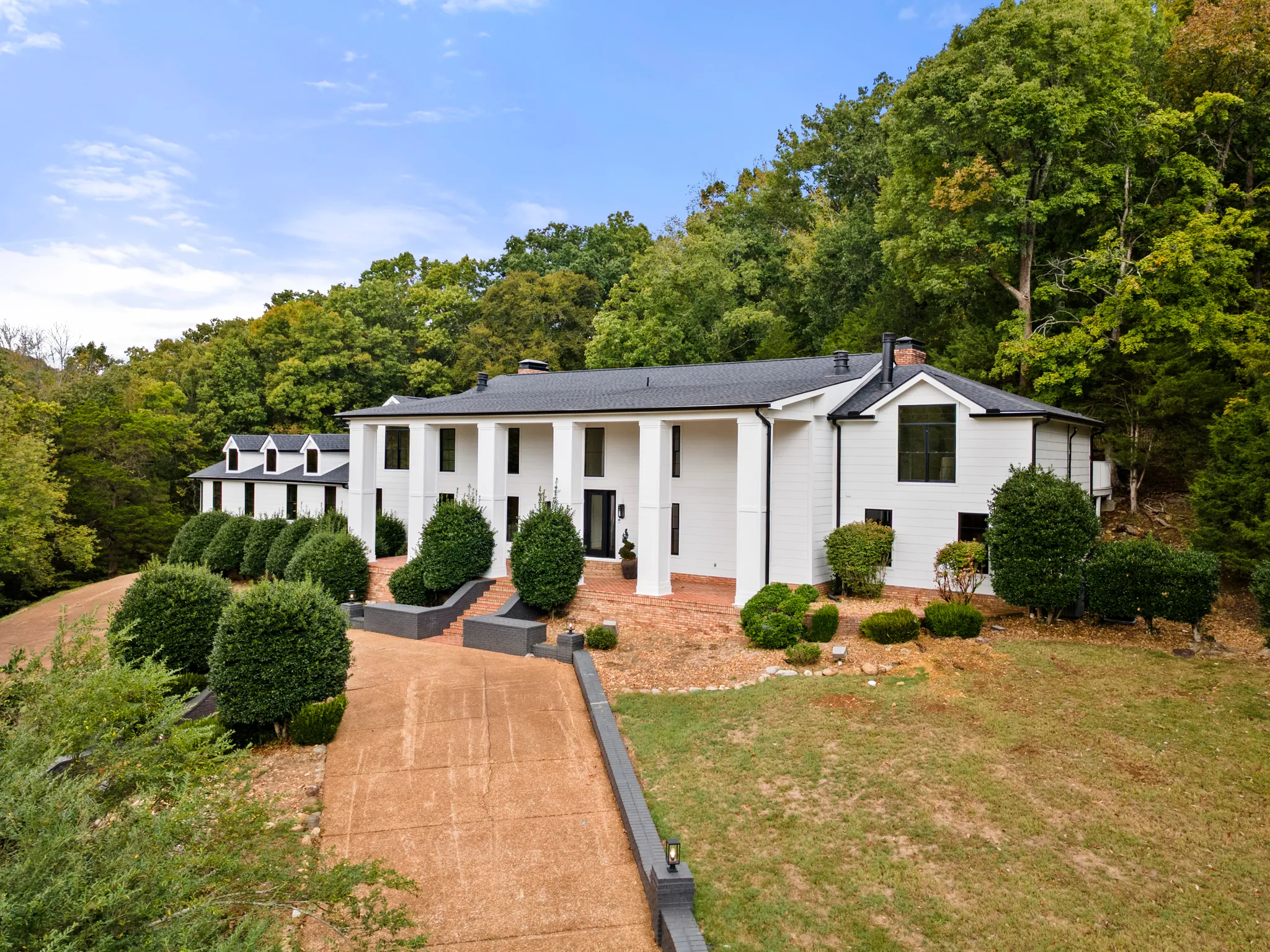 Two-story white home with square porch columns, black shingle roof and dark windows, set on a wooded hillside above a concrete driveway