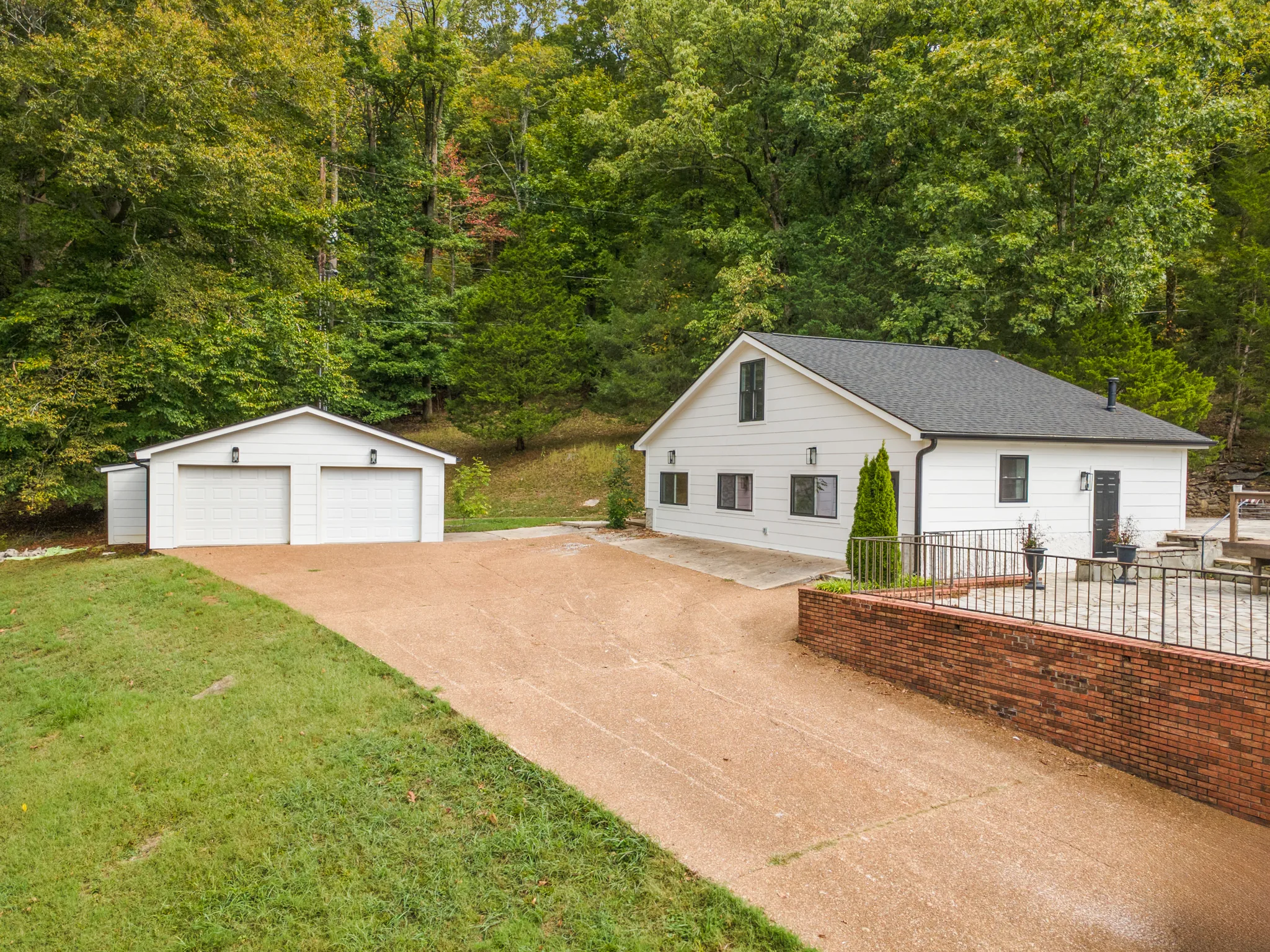 White sided home with dark shingle roof beside a detached two-car garage, wide concrete driveway and brick retaining wall with black metal fence