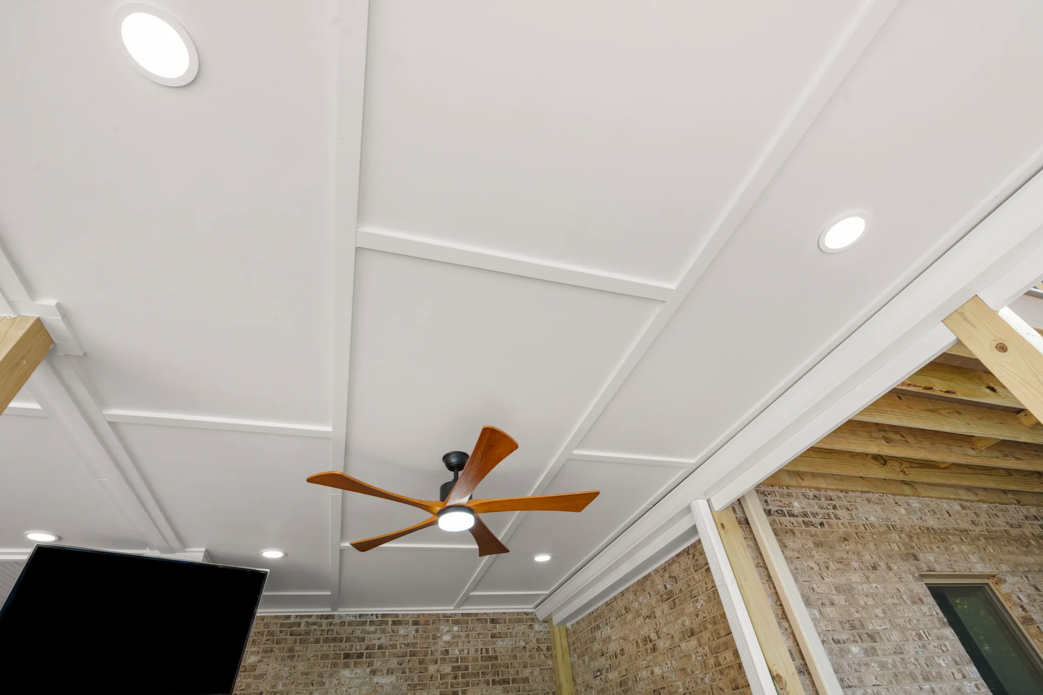 Upward view of a covered porch ceiling with white paneled grid, recessed lights and a five-blade fan with wood blades, adjacent framing under construction