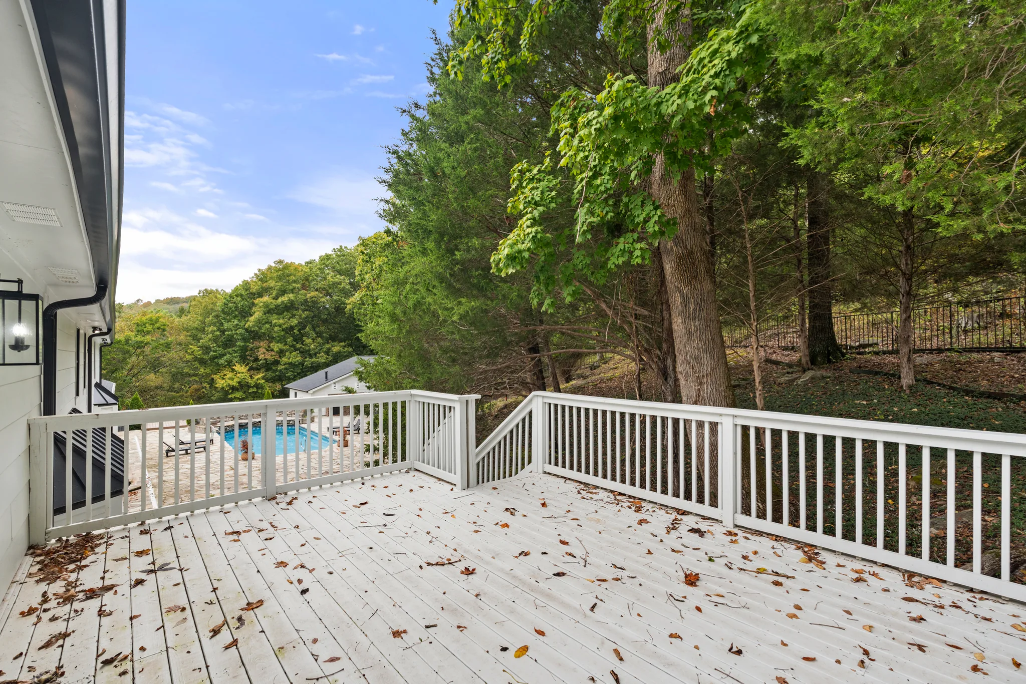 White painted wood deck with white picket-style railing, scattered fallen leaves, and a view downhill to a pool patio and wooded slope