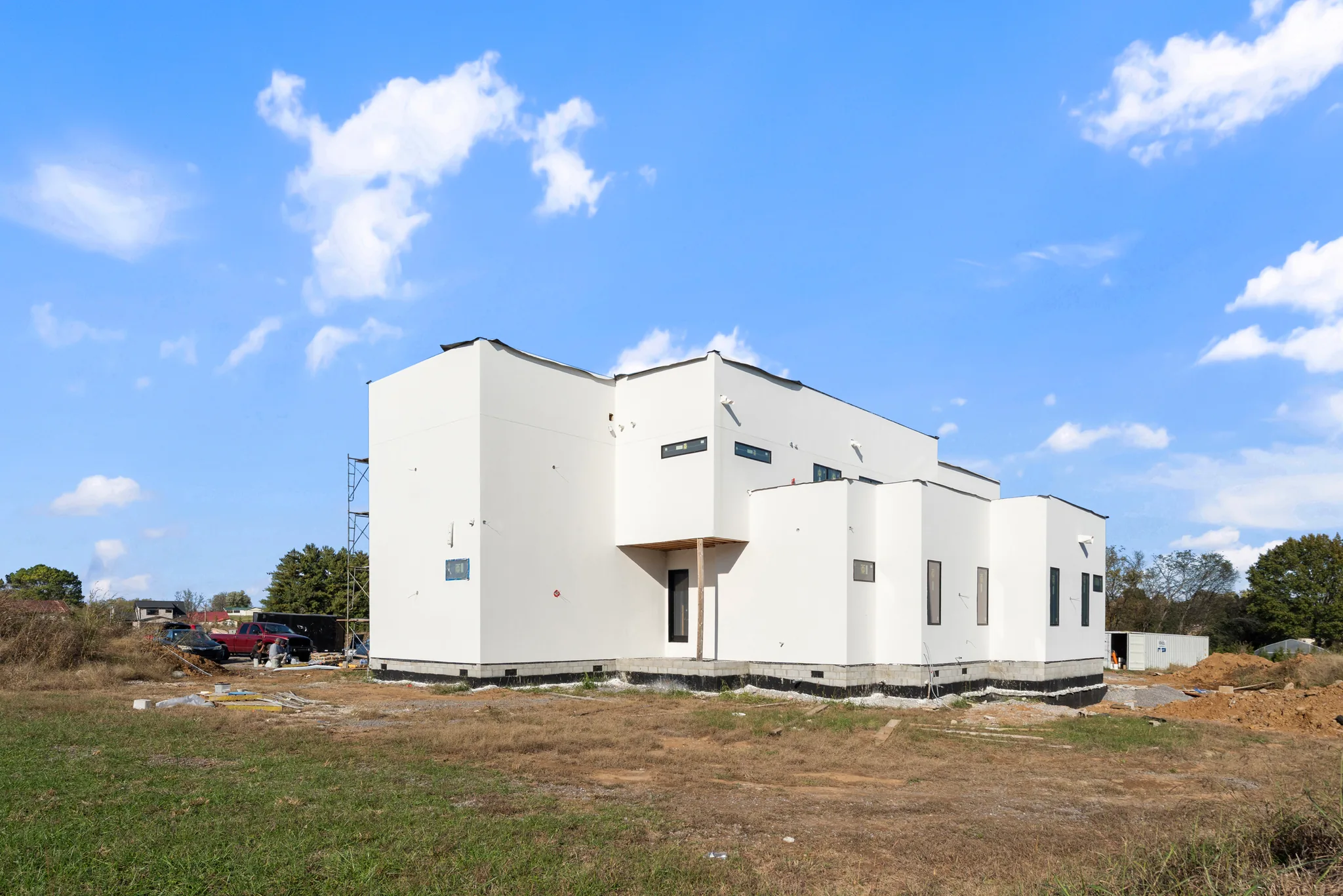 White stucco modern home under construction on a cleared lot with scaffolding, unfinished foundation edges and dark narrow windows under a blue sky