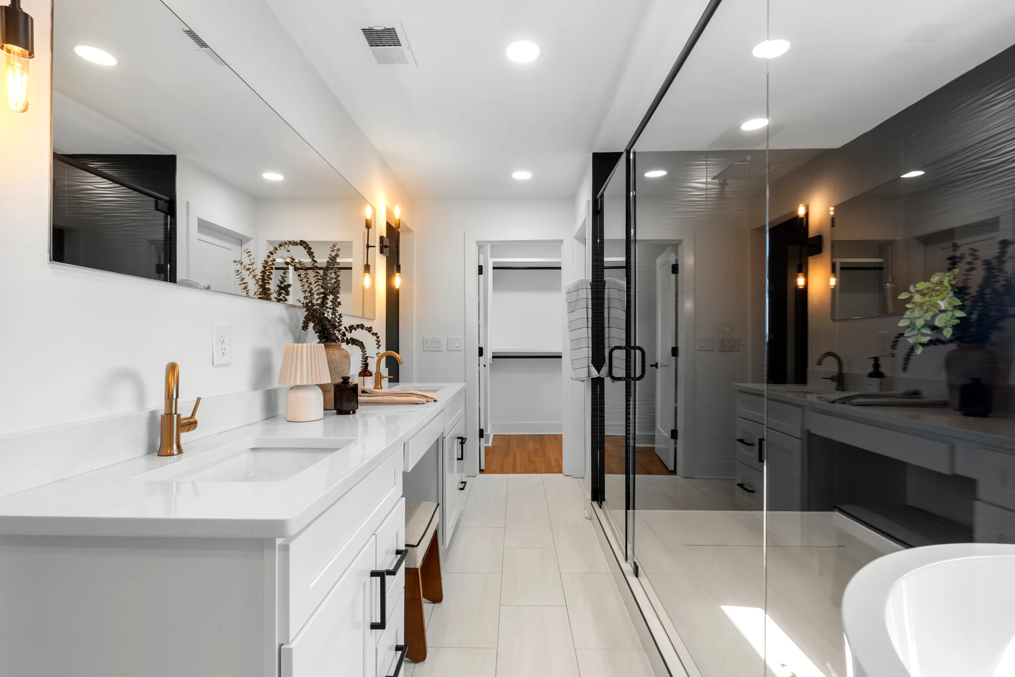 Primary bathroom with a long white quartz vanity, two undermount sinks, brass faucets, frameless glass shower with textured dark tile, and a freestanding tub