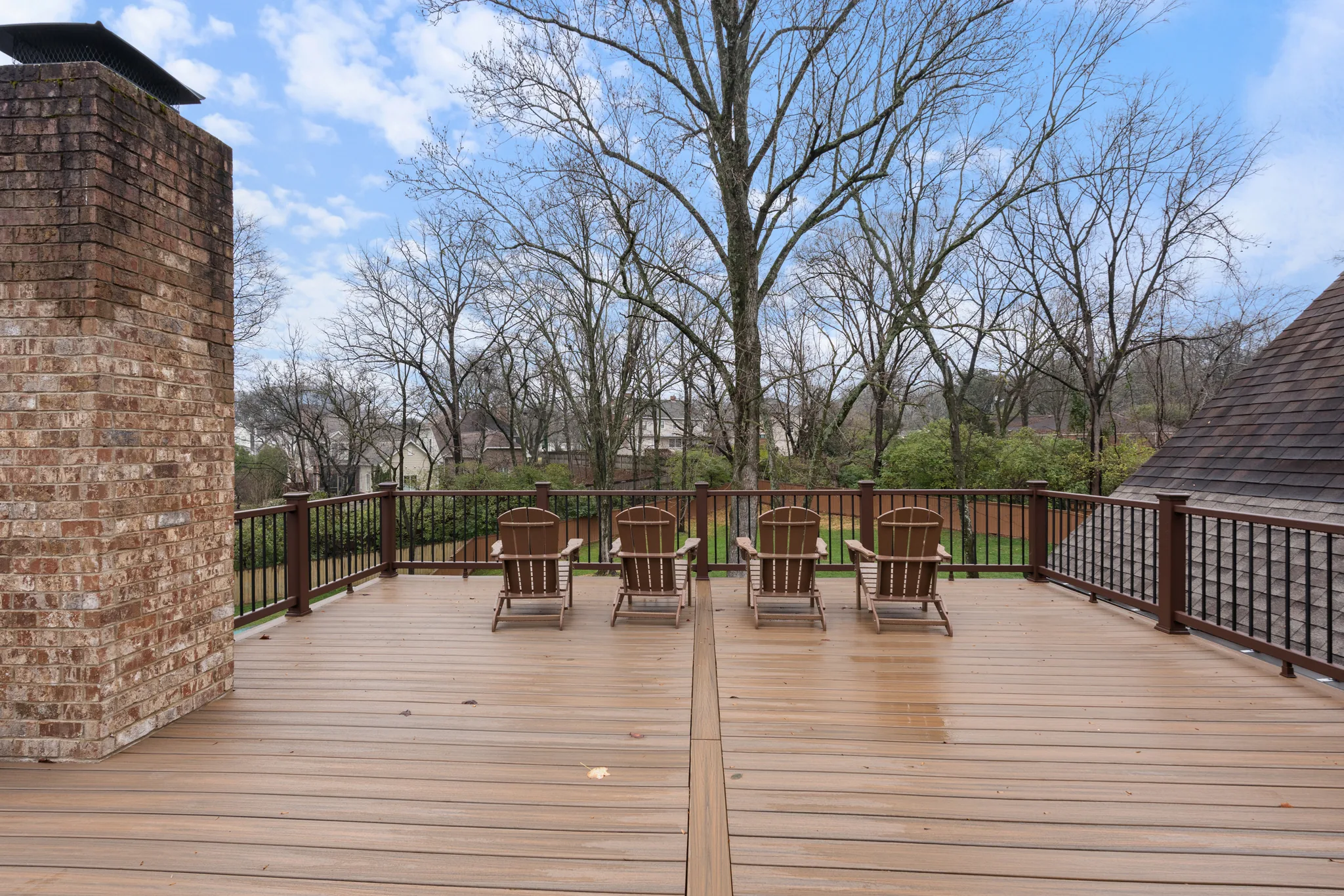 Composite deck with four wood Adirondack chairs lined along a black metal railing, a tall brick chimney at left and bare trees beyond