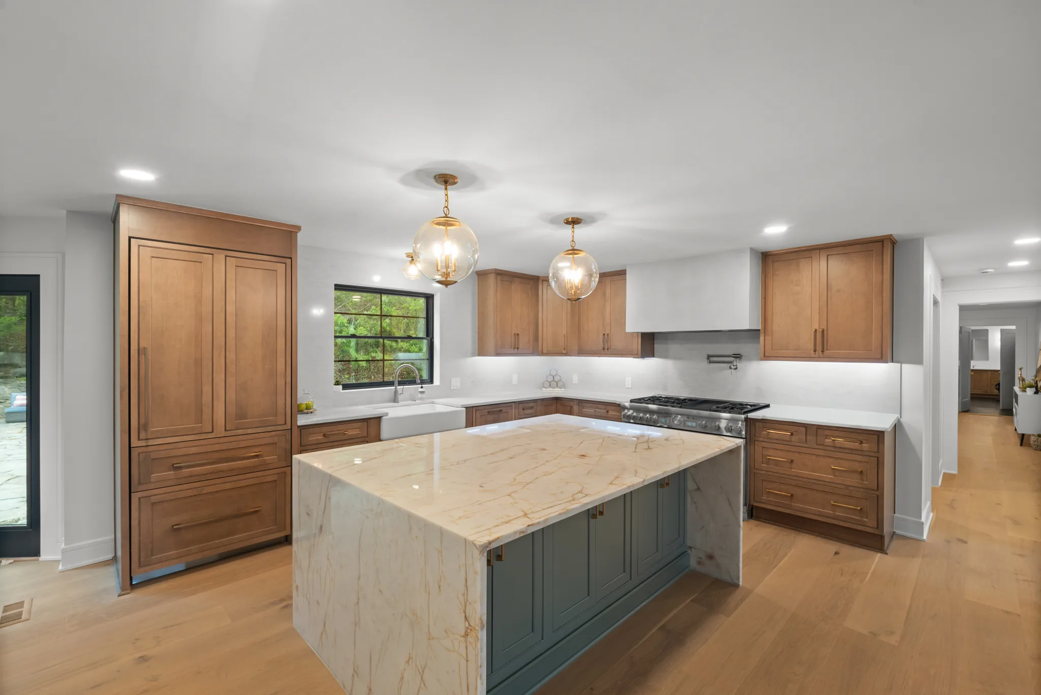 Kitchen with stained wood cabinets, a green island base topped by a veined marble waterfall counter, glass globe pendants and oak floors