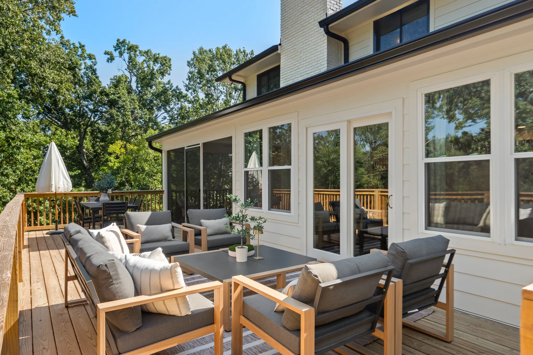 Wood deck with teak lounge chairs and sofa, gray cushions, low table and umbrella, beside a white house with sliding glass doors