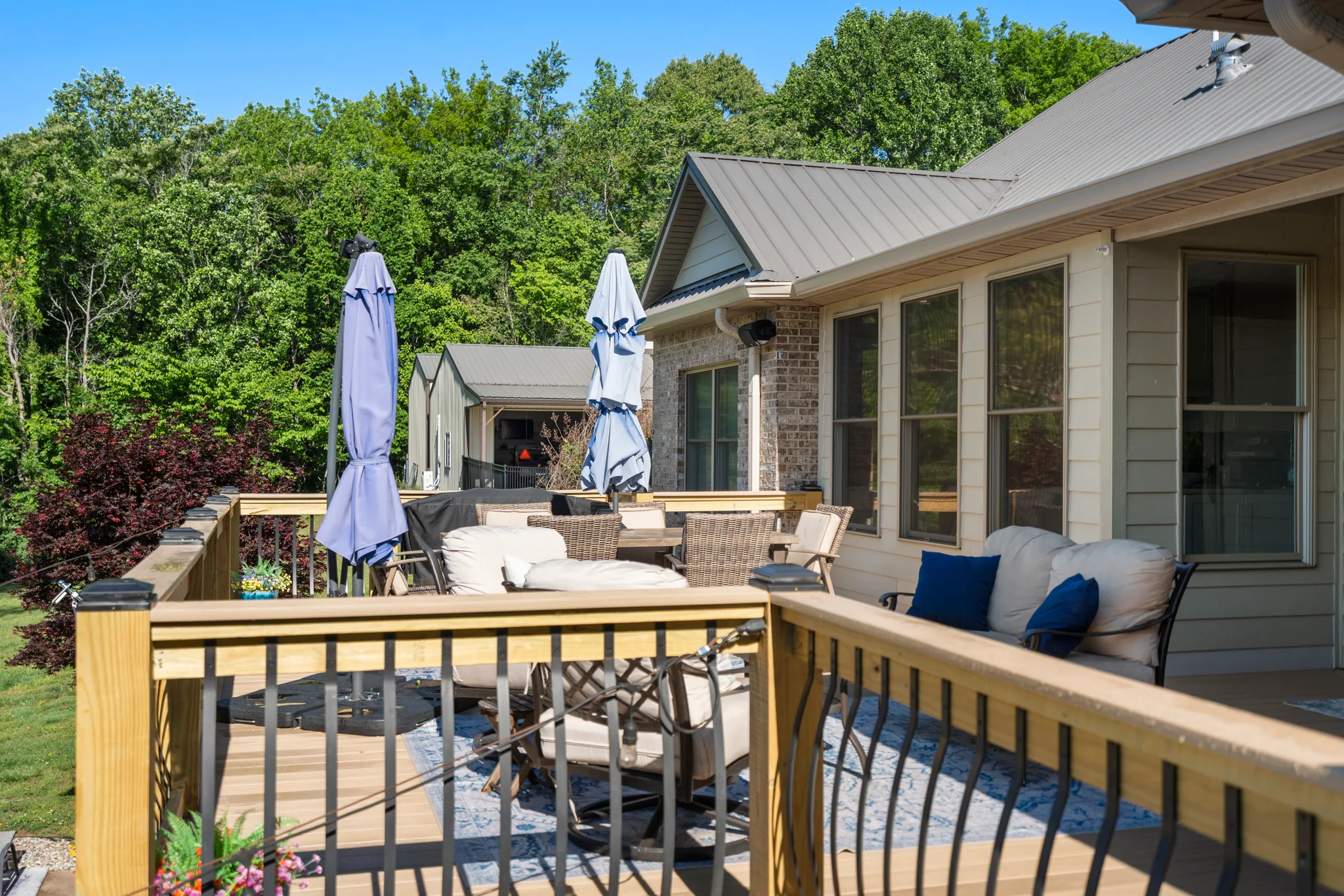 Wood deck with metal baluster railing, wicker seating, closed umbrellas and a patterned outdoor rug beside a brick and siding home