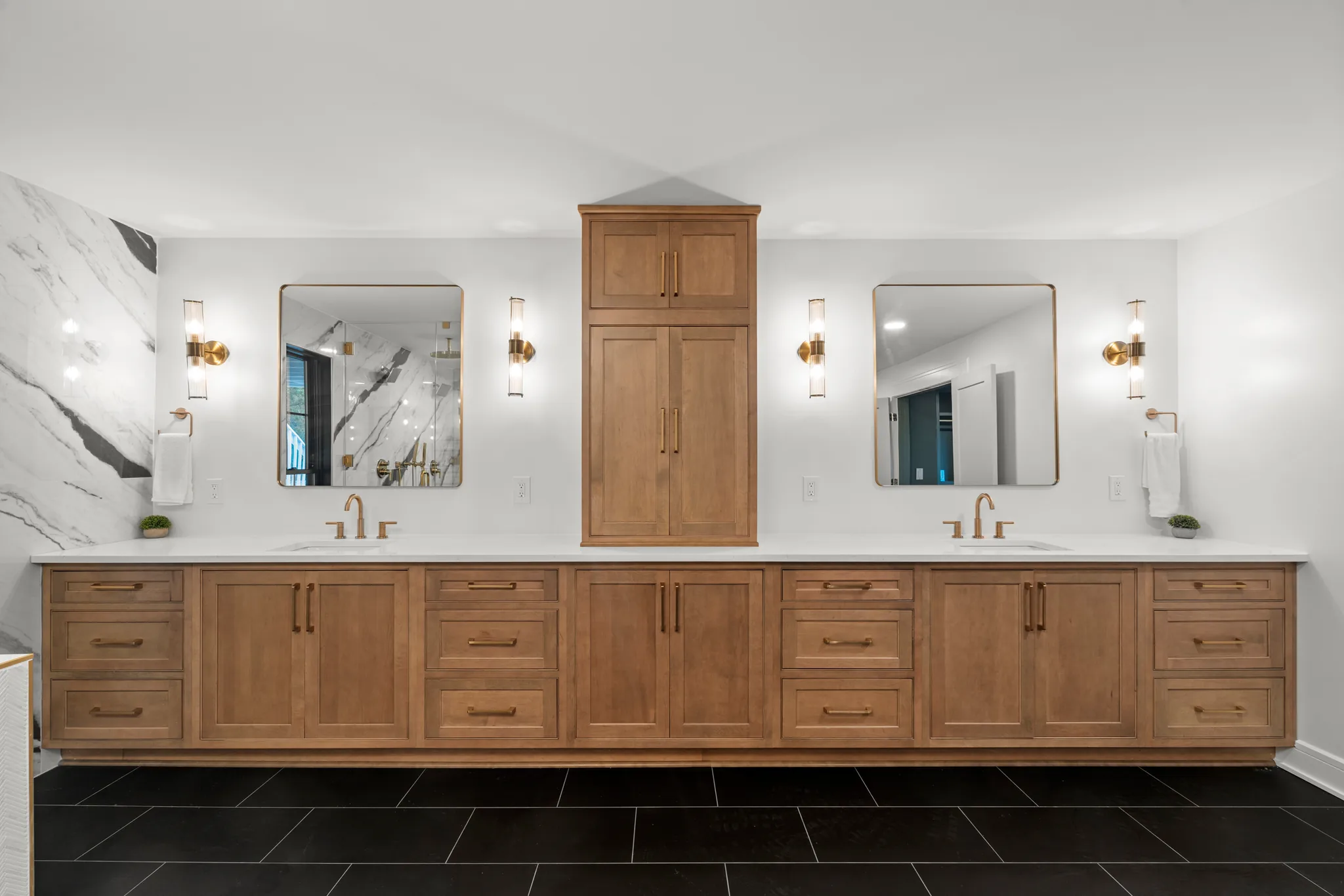 Long stained wood double vanity with white quartz top, center storage tower, brass sconces, two gold-framed mirrors and black tile floor