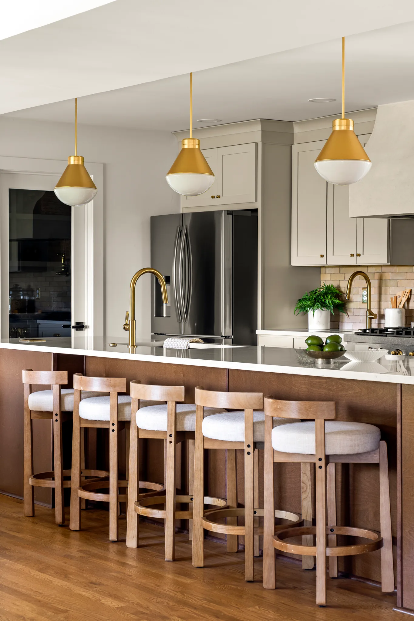 Kitchen island clad in dark wood with white stone counter, five light wood stools, brass pendants and stainless refrigerator