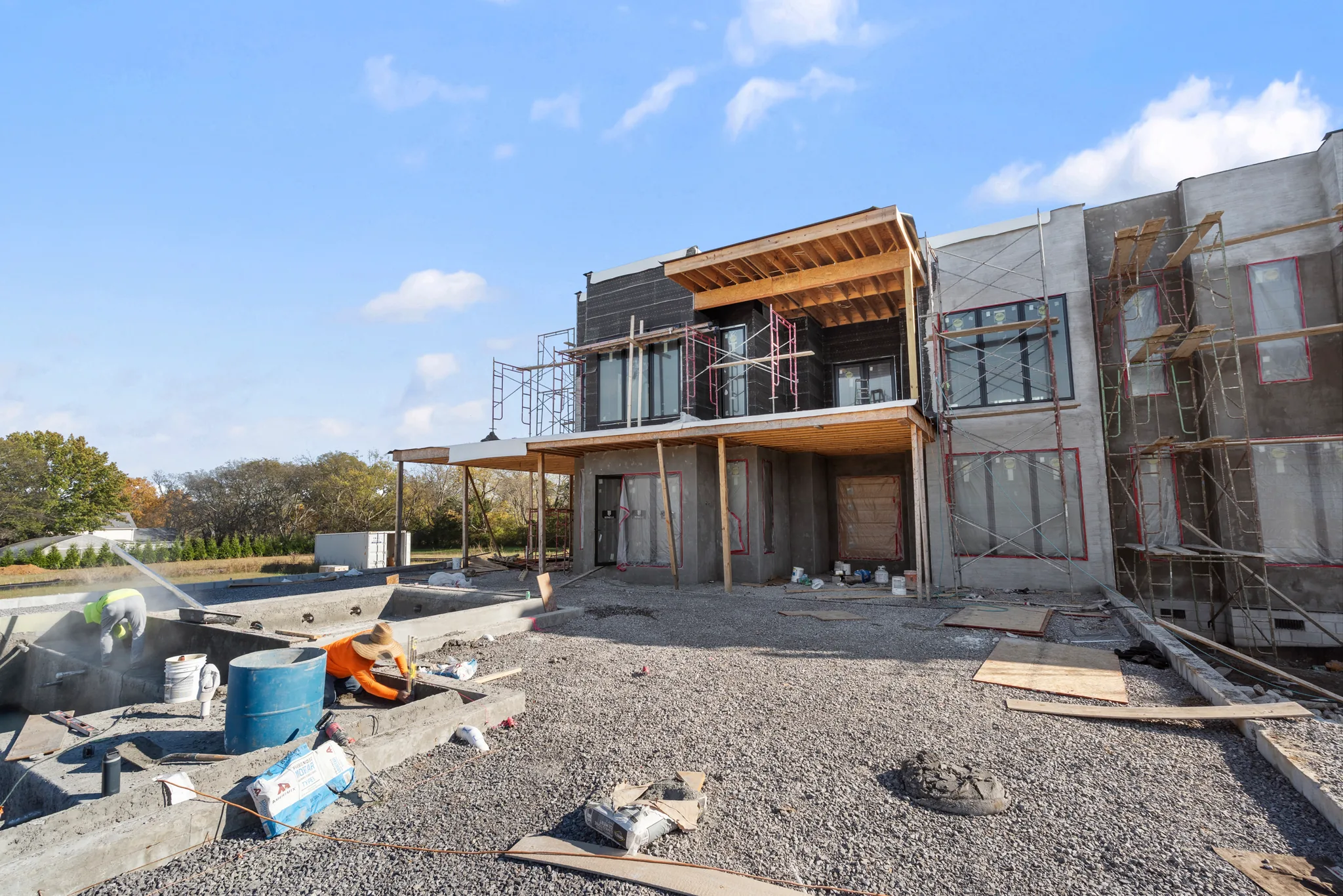 Modern two-story home under construction with scaffolding, dark siding and large windows, workers finishing concrete forms on a gravel yard